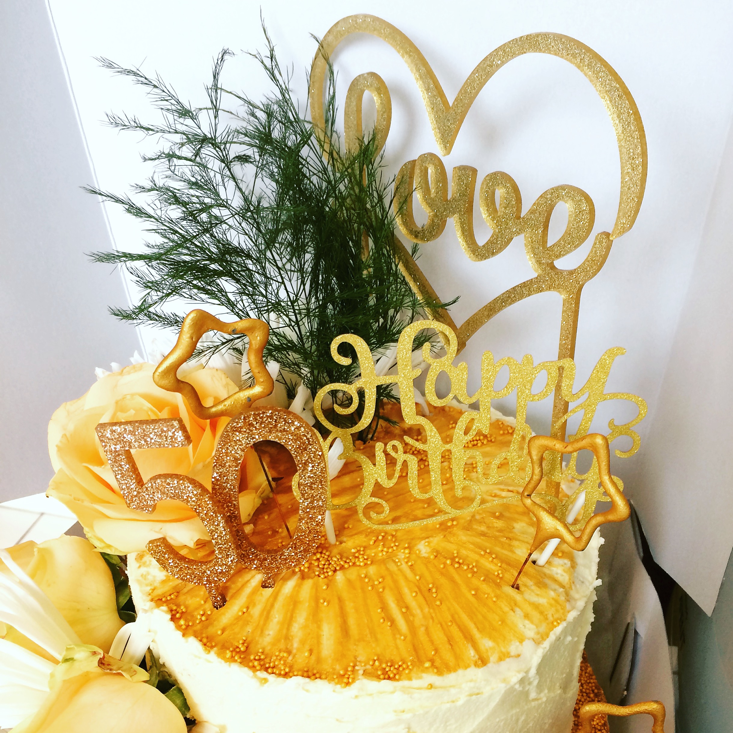 A picture of a 50th birthday cake. It is has vanilla frosting and is decorated with some greenery and words that say Love and Happy Birthday and two golden star shaped candles.
