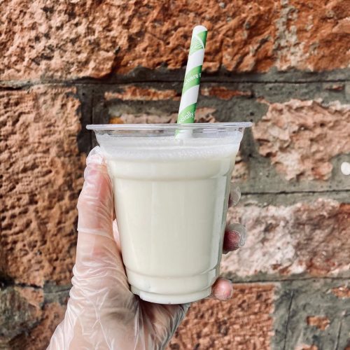 A photo of someone holding a takeaway cup of The Yellow One Smoothie with a green and white stripy straw with an exposed brick wall in the background.