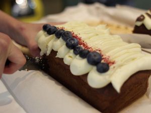 A loaf cake with white frosting and blueberries on top. A person is cutting a slice of it.